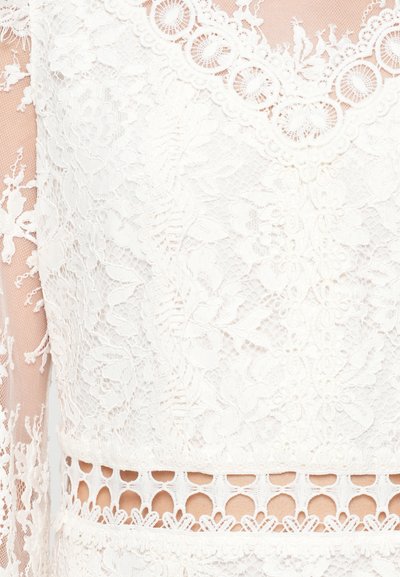 Λευκό φόρεμα από δαντέλα, με περίπλοκα λουλουδάτα μοτίβα, κωνικό λαιμόκομμα και διακοσμητική κοπή στη μέση.