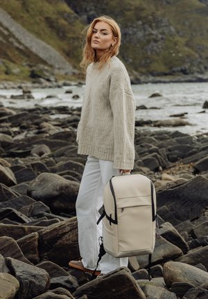 Ung kvinne i beige genser og hvite bukser som holder en beige ryggsekk, stående på en steinete strand med vann og åser i bakgrunnen.