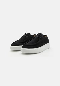 Crne suede cipele sa zaobljenim prstima, bijelim gumenim potplatom i minimalističkim detaljima vezanja. Teksturovan gornji dio s vidljivim šavovima.