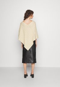 Poncho tricotado em cor creme com um padrão de diamante, sobre uma saia midi de couro preta, usado com sapatos de salto baixo pretos.