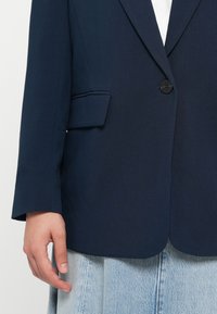 Marineblå blazer med en enkelt sort knap, en brystlomme og en glat stoftekstur. Båret over en lys denimnederdel.