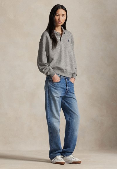 Nainen seisoo kädet taskuissa, päällään harmaa pitkähihainen poolopaita, väljät siniset farkut ja valkoiset tennarit neutraalilla taustalla.