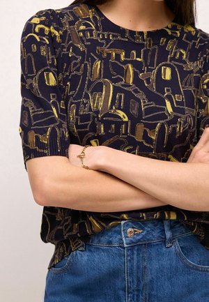Femme portant une chemise bleu marine avec des motifs abstraits de bâtiments dorés et un jean bleu, bras croisés montrant un bracelet doré au poignet.