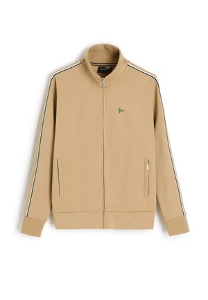 Beżowa bluza sportowa na zamek z wysokim kołnierzem, cienkie czarno-białe paski na rękawach, boczne kieszenie na zamek oraz małe zielone logo na piersi.