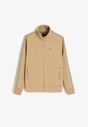 Beżowa bluza sportowa na zamek z wysokim kołnierzem, cienkie czarno-białe paski na rękawach, boczne kieszenie na zamek oraz małe zielone logo na piersi.