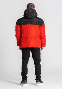 Rød og sort polstret jakke med hætte, der har et syet design og elastiske manchetter, båret over mørke bukser og hvide sneakers.