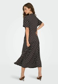Robe midi noire à pois avec manches courtes, une silhouette fluide et des fentes sur les côtés. Tissu lisse avec un léger éclat. Chaussures à talons hauts.