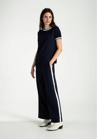 Marineblaues T-Shirt mit weißem Kragen und Ärmelbesatz, kombiniert mit weit geschnittenen marineblauen Hosen mit weißen Seitenstreifen und weißen Knöchelschuhen.
