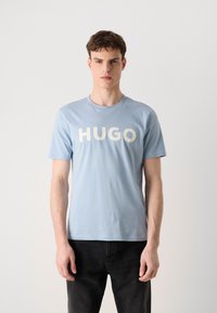 Világoskék pamut póló, kerek nyakkal és merész fehér "HUGO" felirattal az elején. Rövid ujjú, laza szabású, sima textúrával.
