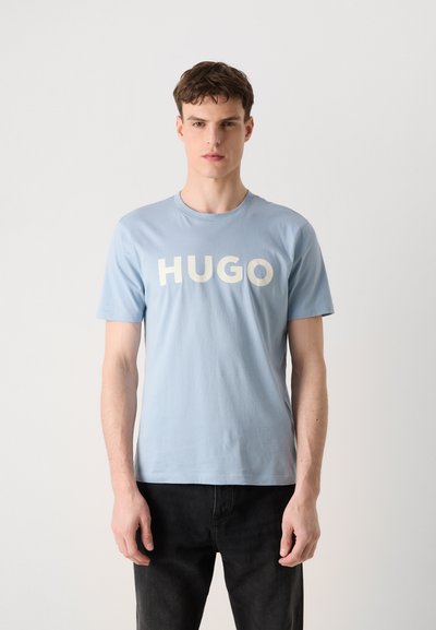 Светлосиня памучна тениска с кръгло деколте и смел бял текст "HUGO" отпред. Къси ръкави, свободна кройка и гладка текстура.