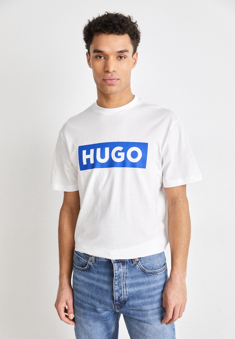 Tricou alb din bumbac cu un grafic rectangular albastru pe care scrie "HUGO." Mânici scurte, guler rotund, croi slim, purtat cu blugi albaștri deschiși.