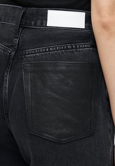 Κοντινό πλάνο μαύρου τζιν από denim με μαύρη δερμάτινη πίσω τσέπη και λευκή ετικέτα επωνυμίας στη μέση, φορεμένο από ένα άτομο.