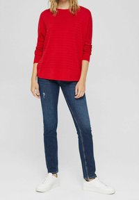 Rød ribbet sweater med lange ærmer, parret med mørke denimjeans og hvide sneakers. Outfittet har en afslappet pasform og en rund halsudskæring.