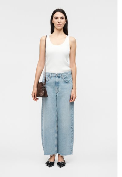 Γυναίκα με λευκό αμάνικο τοπ, ανοιχτά μπλε φαρδιά τζιν, μαύρα μυτερά φλατ παπούτσια και μικρή καφέ τσάντα χιαστί.