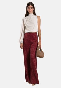 Blouse blanche drapée sur une épaule avec une texture douce, associée à un pantalon large bordeaux avec un motif subtil, tenant un petit sac beige.