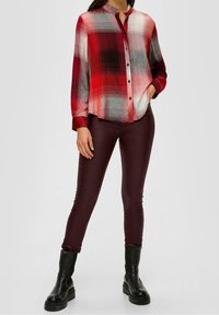 Camisa a cuadros en rojo y gris con botón en la parte delantera, mangas largas y cuello mao, combinada con pantalones ajustados en burdeos oscuro y botas negras.