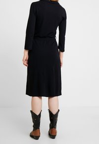 Musta mekko, jossa on pitkät hihat, kaulus ja solmittava vyötärö. Alaosa on sileä. Yhdistetty mustiin ja ruskeisiin lännen bootseihin.