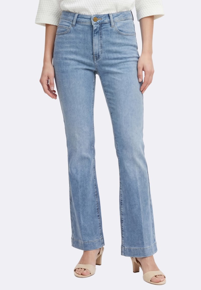 Femme portant un jean droit bleu clair, un haut blanc texturé à manches longues et des sandales beiges à talons.
