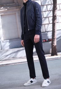 Marinblå bomberjacka med dragkedjeficka, svart turtleneck, mörka byxor och vita sneakers med marinblå detaljer. Släta, texturerade material.