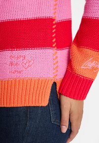 Eine Hand ruht an der Hüfte und trägt einen gestreiften Strickpullover in pink, rot und orange mit bestickten Herzen und Text, kombiniert mit dunkelbblauen Jeans.