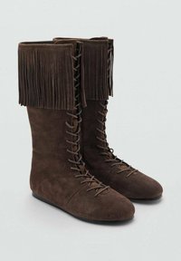 Botas marrones de ante hasta la rodilla con un puño con flecos, presentan un diseño de cordones en el frente y suelas planas. Textura suave y acabado liso.