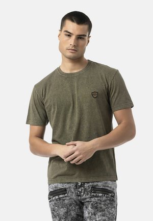 Jonge man met kort haar die een olijfgroen T-shirt en acid-wash grijze spijkerbroek met voorvakken met rits draagt, staand tegen een witte achtergrond.