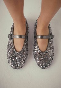Silberne, mit Pailletten besetzte Mary Jane Schuhe mit einer runden Zehenpartie, flacher Sohle und einem Schnallenriemen um den Knöchel. Mit überlappenden Pailletten strukturiert.