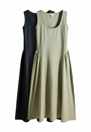 Frau in einem langen grauen Kleid steht neben einem runden Pool, umgeben von grünen Hecken und Laub in einem Garten im Freien.