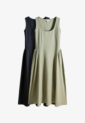 Femme en longue robe grise debout à côté d'une piscine circulaire, entourée de haies vertes et de feuillage dans un jardin extérieur.