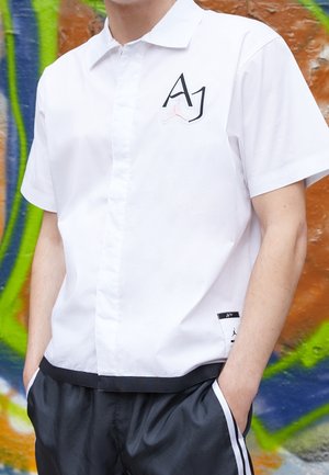 Person iført en hvid t-shirt med korte ærmer med "AJ" logo og sort kant, stående mod en farverig graffiti-væg, hænder i lommerne på sorte bukser.