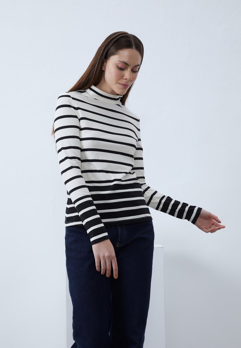 Haut à manches longues rayé noir et blanc avec un col montant et une coupe ajustée. Associé à un jeans bleu foncé, présenté sur un fond clair.