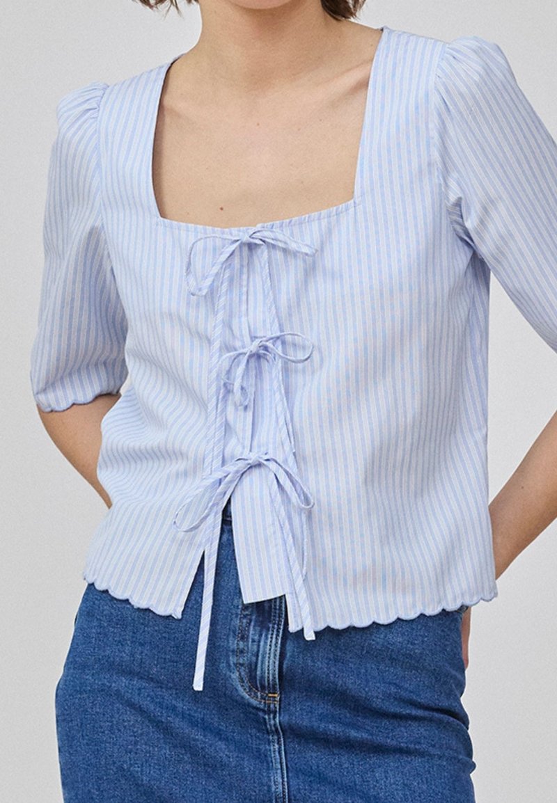 Femme portant un chemisier rayé bleu clair et blanc avec un col carré et trois nœuds noués sur le devant, associé à une jupe en jean.