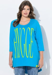 Türkises, übergroßes T-Shirt mit einem auffälligen, leuchtend gelben "HYGGE"-Grafik, Rundhalsausschnitt, 3/4-Ärmeln und einem abgerundeten Saum.