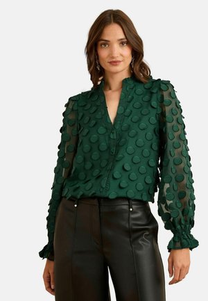 Blusa verde a pois con maniche trasparenti, frontale con bottoni e polsini arricciati. Abbinata a pantaloni in pelle nera.