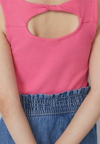 Vrouw in een roze mouwloze ribbeltrui met een uitsnede aan de voorkant en een hoge blauwe elastische spijkerrok, met gekruiste armen.