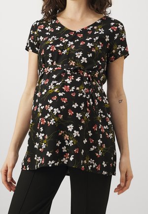Zwangere vrouw draagt een zwarte bloemen top met korte mouwen en strikdetail, gecombineerd met een zwarte broek.
