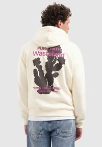 Persoon in een crèmekleurige hoodie met zwarte cactusgrafiek en roze tekst "Pure Path Wasteland Own the Journey Arizona Utah Nevada" op de achterkant.