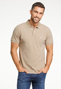 Beige pikétröja med korta ärmar, treknappsstängning och en liten vit logotyp på vänster bröst. Buren med blå jeans.