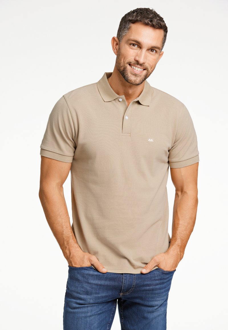 Beige pikétröja med korta ärmar, treknappsstängning och en liten vit logotyp på vänster bröst. Buren med blå jeans.