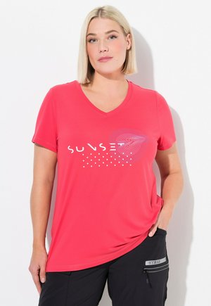 Lyshåret kvinde i en rød T-shirt med V-hals med teksten "SUNSET" og et abstrakt design, iført sorte bukser med en lynlåslomme.