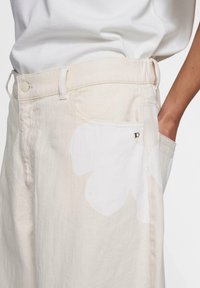 Pantalones de mezclilla beige claro con un gran patrón floral blanco en la parte frontal. Cuenta con un bolsillo lateral y un botón de metal en la cintura.