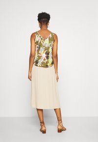 Top in tessuto leggero color oliva con fantasie floreali in rosa, giallo e bianco, abbinato a una gonna beige corta fino al ginocchio e sandali con cinturini marroni.