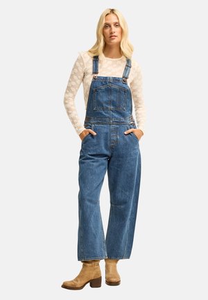 Femme aux cheveux blonds portant un haut transparent à manches longues fleuri sous une salopette en jean bleu et des bottines beige, debout les mains dans les poches.