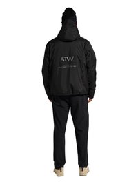 Person trägt schwarze Kapuzenjacke mit „ATW“- und Textlogo auf dem Rücken, schwarze Hose, schwarze Mütze und beigen Schuhe, blickt auf weißem Hintergrund nach hinten.