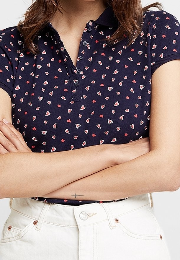 Mörkblå polo med små röda och vita hjärtmönster, korta ärmar och krage, tillverkad av mjukt bomullsmaterial.