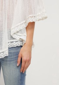Biały dzianinowy top z delikatnymi koronkowymi akcentami wzdłuż rękawów i dołu, w połączeniu z niebieskimi dżinsami. Miękka tekstura, luźny krój.