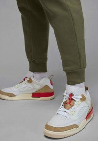 Persona che indossa sneakers alte bianche, beige e rosse con calzini bianchi e pantaloni jogger verde oliva su sfondo grigio.