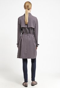 Szary płaszcz trench z luźnym krojem, detalami paska, peleryną z tyłu i długimi rękawami, w połączeniu z ciemnymi dżinsami i szarymi butami.