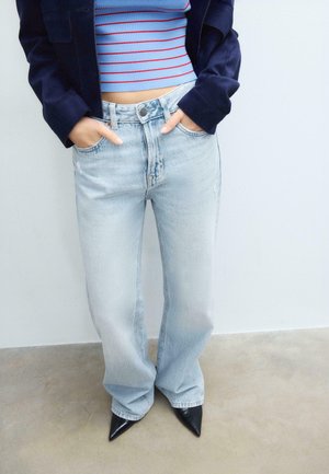 Personne portant un jean large bleu clair, des chaussures pointues noires, un haut court rayé bleu et une veste bleu foncé avec les mains dans les poches.