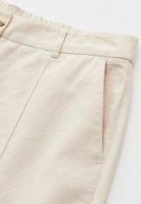 Détail d'un pantalon beige clair avec ceinture, passants de ceinture, poche latérale et fermeture à bouton visible sur un tissu texturé.